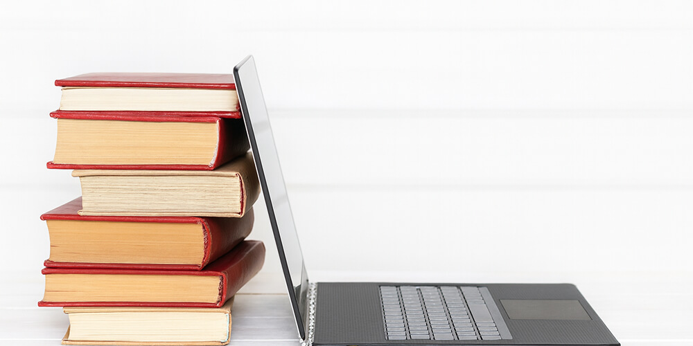 Pile de livres reliés à côté d’un ordinateur portable ouvert sur fond blanc, symbolisant la rédaction d’un mémoire entre supports papier et numérique.