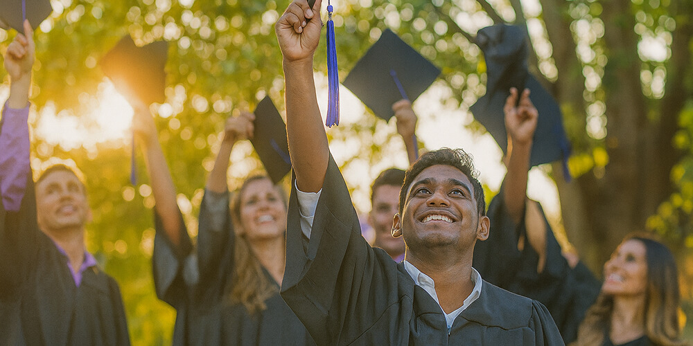 Jeunes diplômés souriants en tenue de graduation levant leur toque universitaire lors d’une cérémonie en plein air au coucher du soleil.