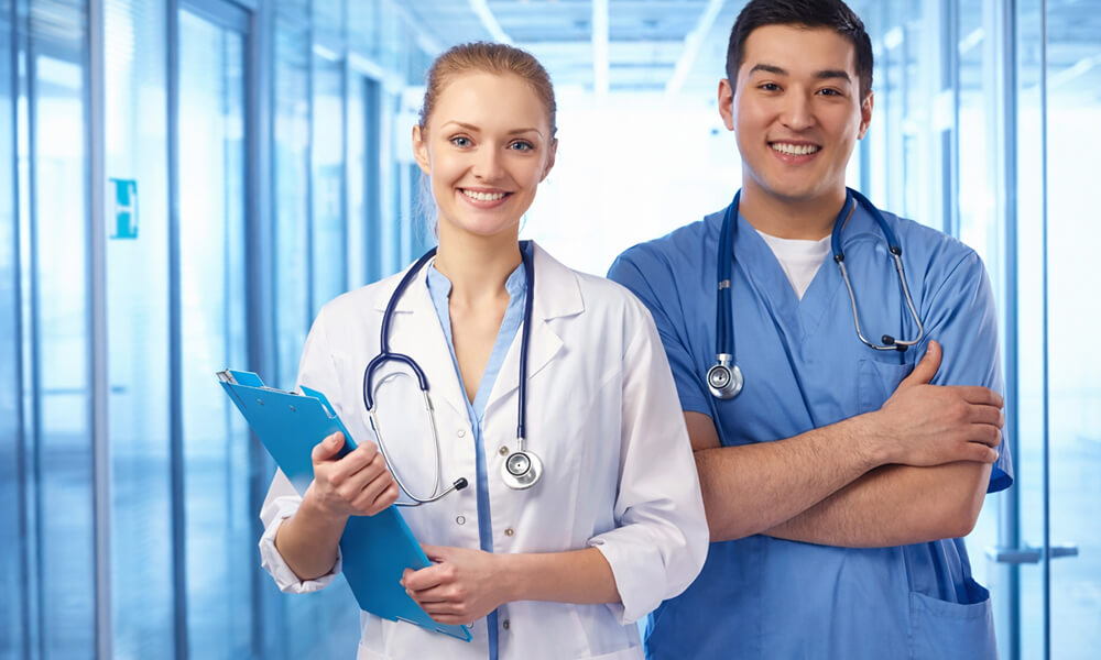 Deux professionnels de santé souriants – une médecin en blouse blanche avec stéthoscope et clipboard, et un infirmier en tenue médicale bleue – debout dans un couloir d’hôpital moderne et lumineux.