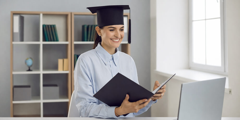 Étudiante diplômée en toge et mortier souriant devant un ordinateur portable, tenant un dossier
