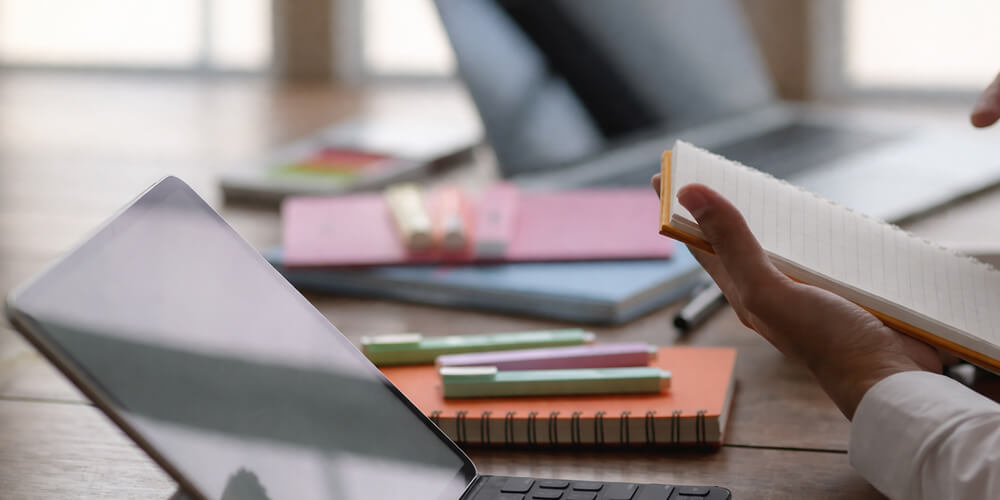 Étudiant consultant un ordinateur portable et prenant des notes dans un carnet, avec fournitures scolaires sur un bureau.
