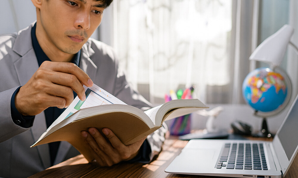 Homme en train de préparer son mémoire d’expertise comptable, consultant un livre et des documents à côté d’un ordinateur portable.