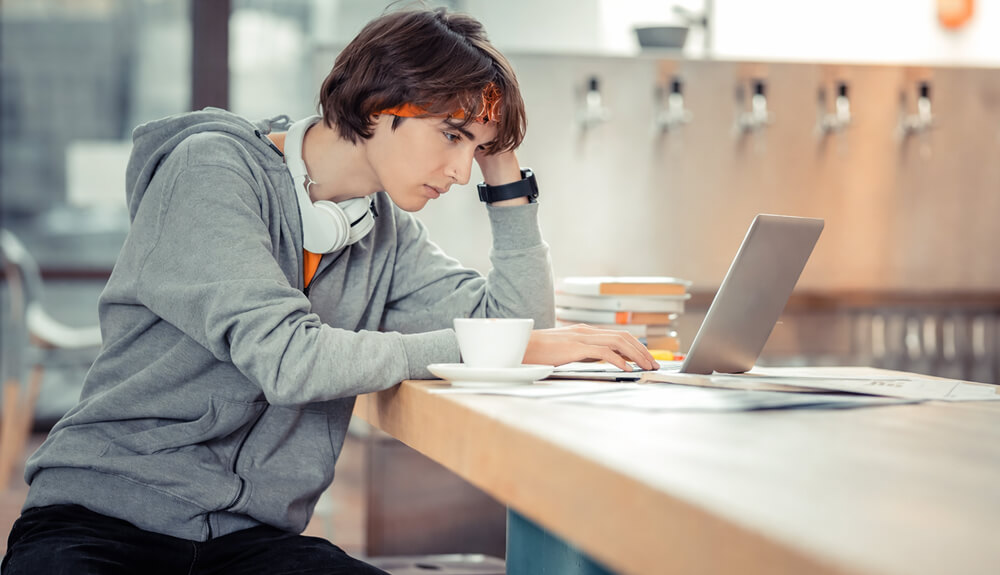 Étudiant concentré corrigeant son mémoire sur un ordinateur portable dans un espace de travail, avec livres et café à proximité.