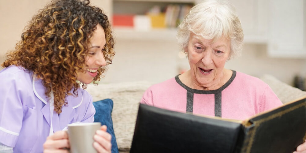 Aide-soignante et femme âgée regardant un album photo sur un canapé, dans un salon lumineux