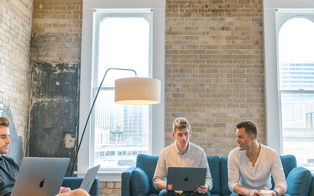 Trois hommes collaborant dans un bureau de style industriel, utilisant des ordinateurs portables pour une réunion informelle.