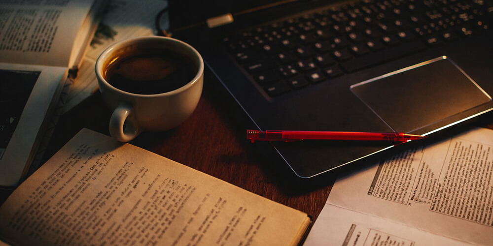 Espace de travail académique avec ordinateur portable, livres ouverts, tasse de café et documents de recherche, illustrant la rédaction d’un mémoire universitaire.
