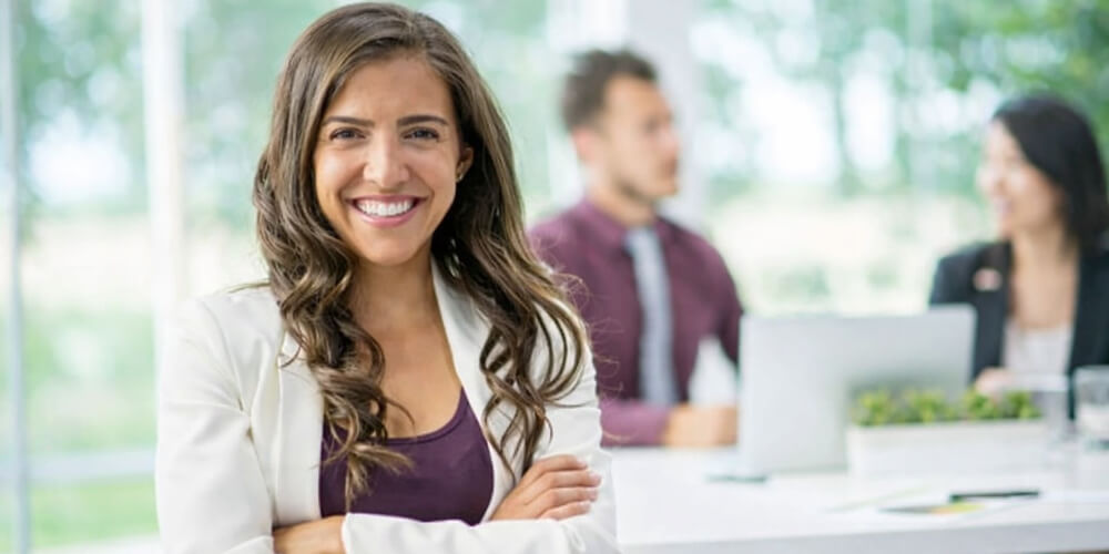 Femme souriante en tenue professionnelle, debout dans un bureau moderne avec une équipe en arrière-plan.