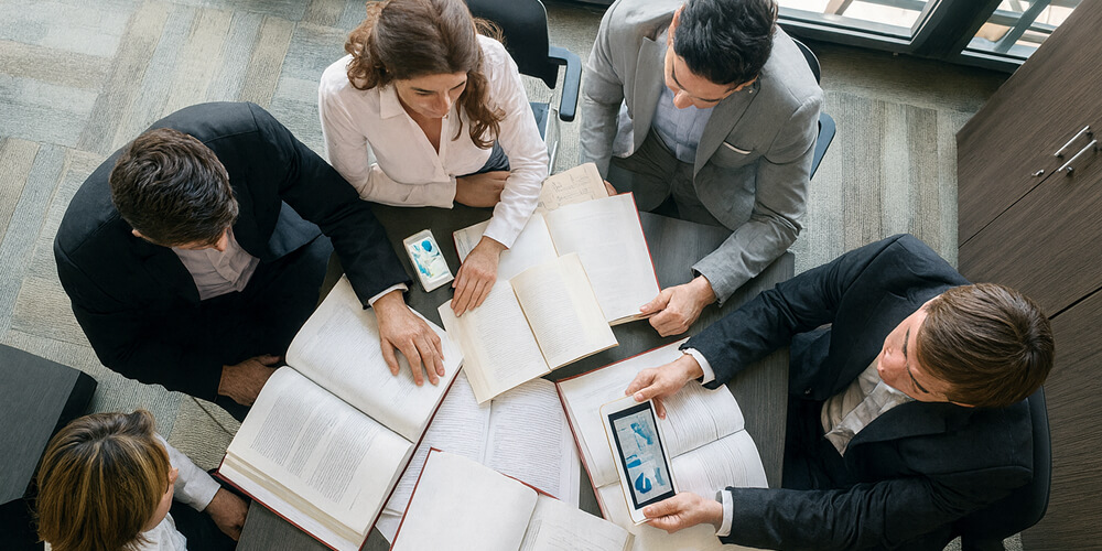 Groupe de professionnels en réunion autour de livres et documents, collaborant sur un projet de recherche avec tablettes et smartphones, vue aérienne dans un bureau moderne.