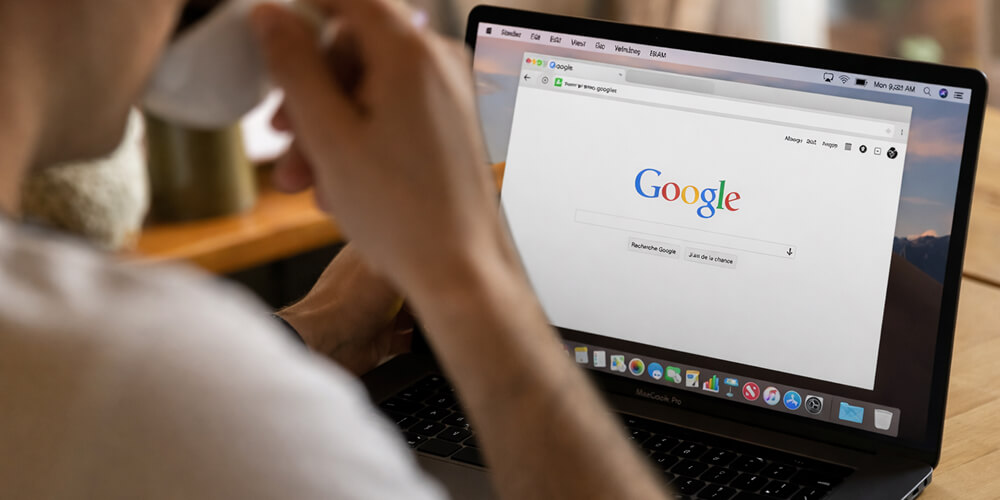 Personne travaillant sur un ordinateur portable affichant la page d’accueil Google, assise à une table en bois et buvant un café dans un environnement calme et lumineux.