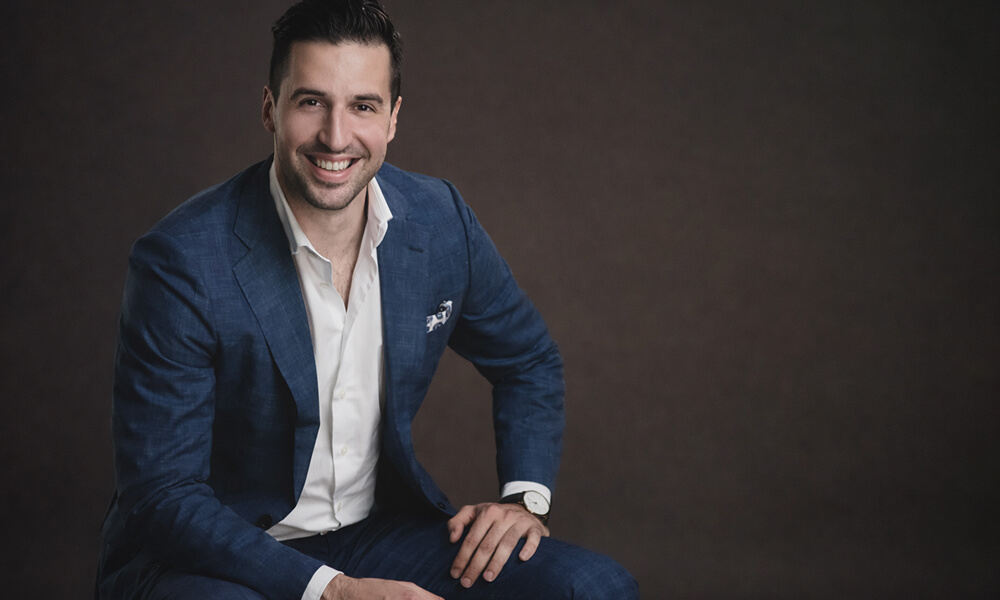 Portrait professionnel d’un homme souriant en costume bleu, assis, sur fond sombre, style corporate.