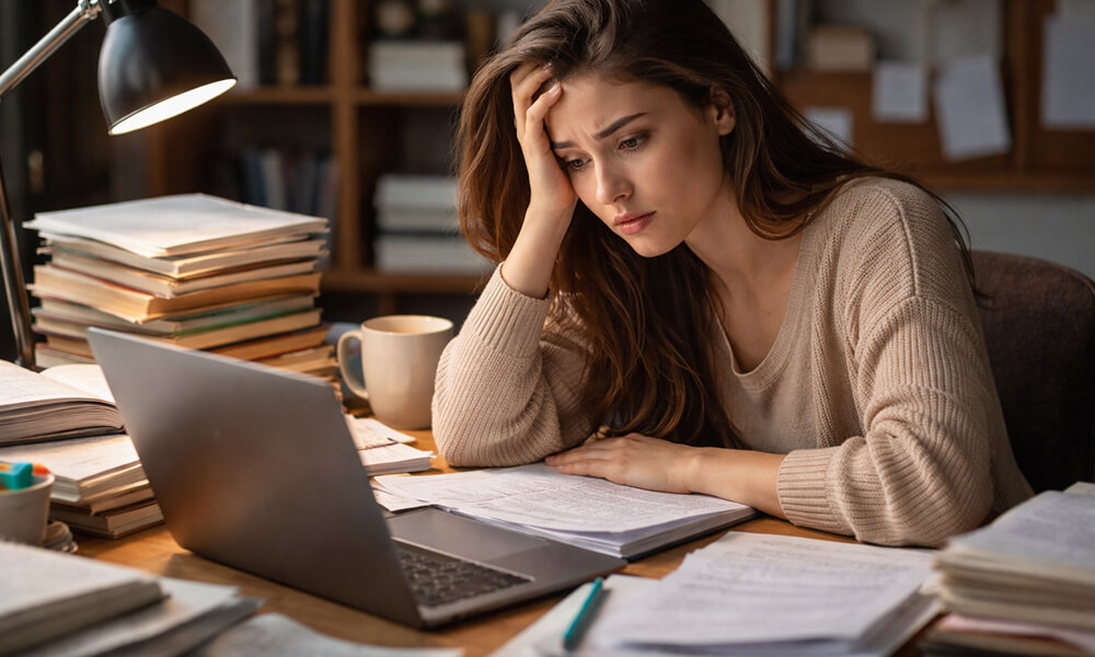 Étudiante stressée assise à un bureau encombré de livres et de feuilles, se tenant la tête devant son ordinateur portable.