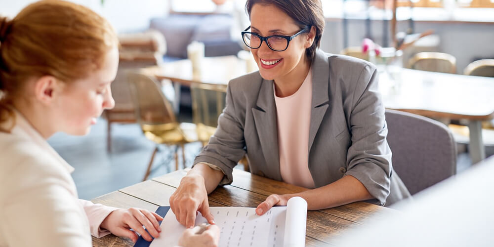 Deux femmes en réunion professionnelle, discutant d’un document sur une table en bois dans un espace de travail lumineux.