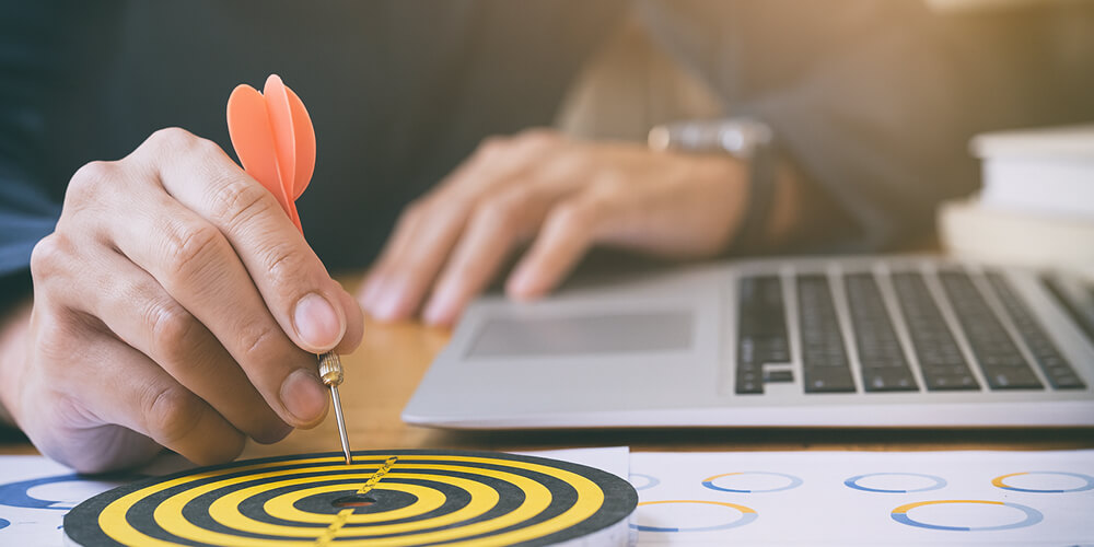 Entrepreneur plaçant une fléchette au centre d’une cible, symbole d’objectifs commerciaux atteints sur un bureau avec ordinateur et graphiques.