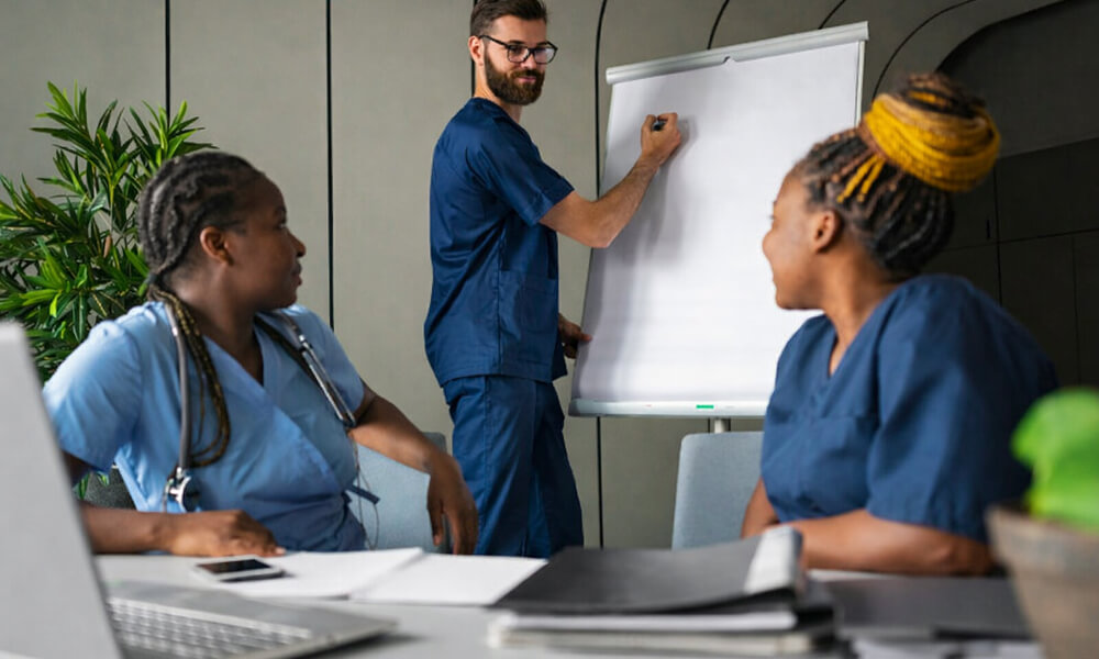 Réunion d’équipe soignante avec infirmiers en tenue médicale échangeant autour d’un tableau blanc lors d’une session de formation ou de coordination.