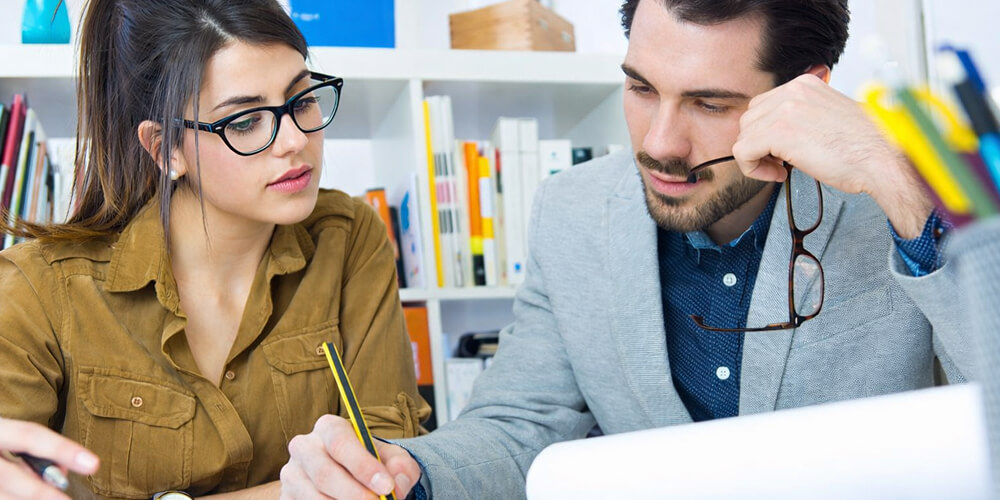 Étudiant et étudiante travaillant ensemble sur la rédaction d’un mémoire universitaire, échangeant autour d’un document académique dans un bureau.