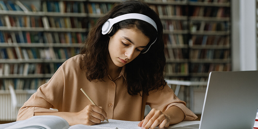 Étudiante studieuse en bibliothèque, portant un casque audio et prenant des notes tout en utilisant un ordinateur portable pour ses recherches académiques.